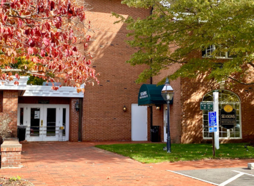 Brick building entrance with a green awning, surrounded by trees and autumn leaves, featuring a lamp post and signage.