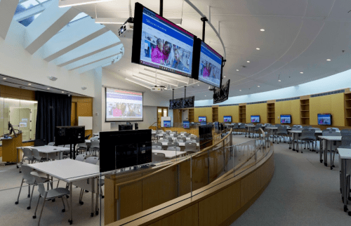 A modern classroom with multiple screens, tables, and chairs, featuring large windows and a bright, open design.