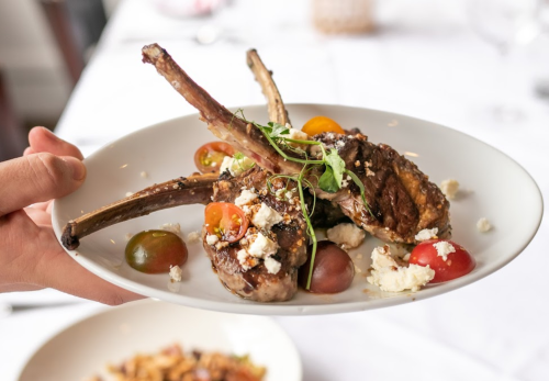 A hand holds a plate with grilled lamb chops, cherry tomatoes, and crumbled cheese, garnished with microgreens.