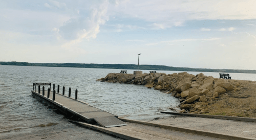 A wooden dock extends into a calm lake, with rocky shorelines and benches in the background under a cloudy sky.