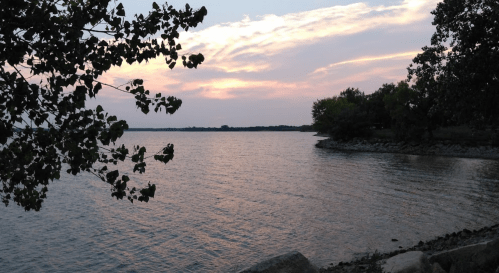 A serene lakeside view at sunset, with gentle waves and trees framing the scene against a colorful sky.