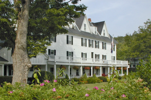 A large white building with multiple balconies, surrounded by greenery and flowers, under a clear blue sky.