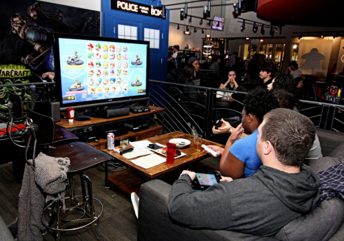 Group of people playing video games on a large screen in a lively gaming venue, with snacks and drinks nearby.