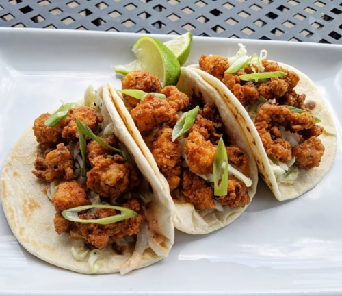 Three crispy fried chicken tacos topped with green onions, served with lime wedges on a white plate.