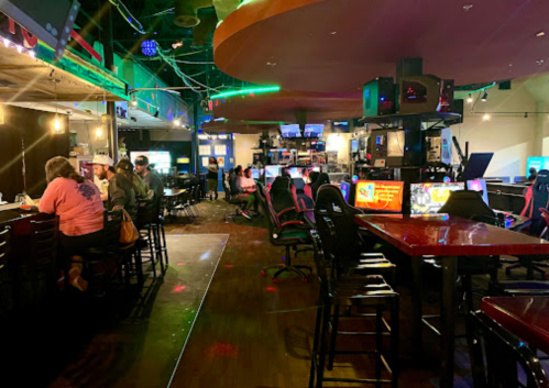 A lively gaming lounge with patrons at tables and gaming stations, featuring colorful lights and a vibrant atmosphere.