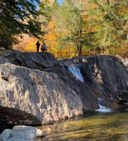 Two people stand on a rocky ledge near a small waterfall, surrounded by autumn foliage and trees.
