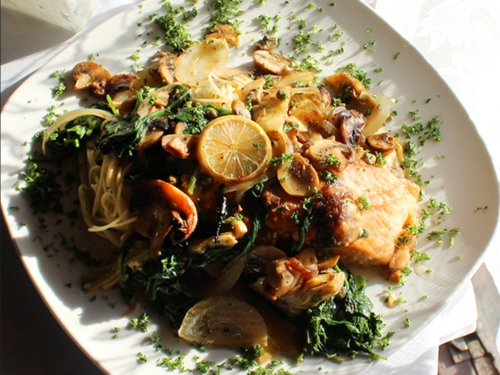 A plate of pasta topped with sautéed vegetables, lemon slices, and a piece of fish, garnished with herbs.