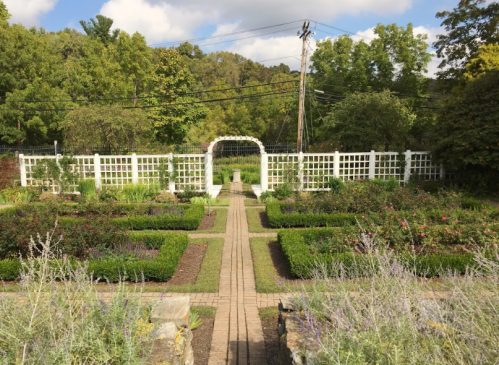 A serene garden with a pathway leading to a white trellis, surrounded by lush greenery and neatly arranged flower beds.
