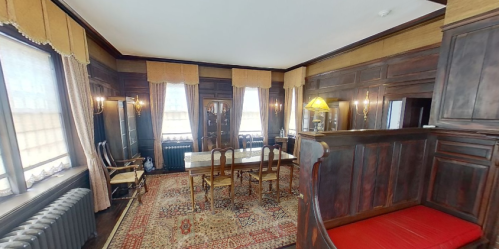 A cozy, vintage room with wooden walls, a dining table, chairs, and a patterned rug, illuminated by soft lighting.