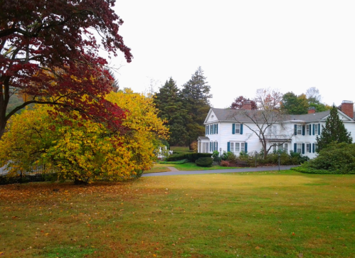 A white house surrounded by colorful autumn trees and a grassy lawn on a cloudy day.