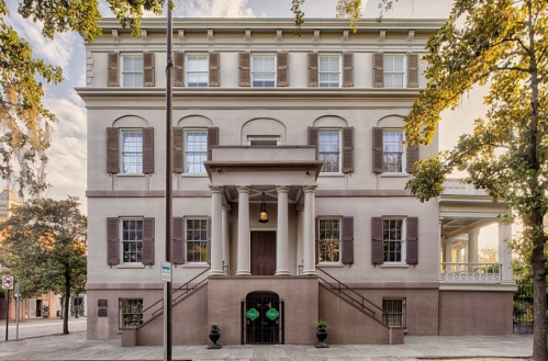 A historic building with a grand entrance, large windows, and a staircase, surrounded by trees and a street sign.