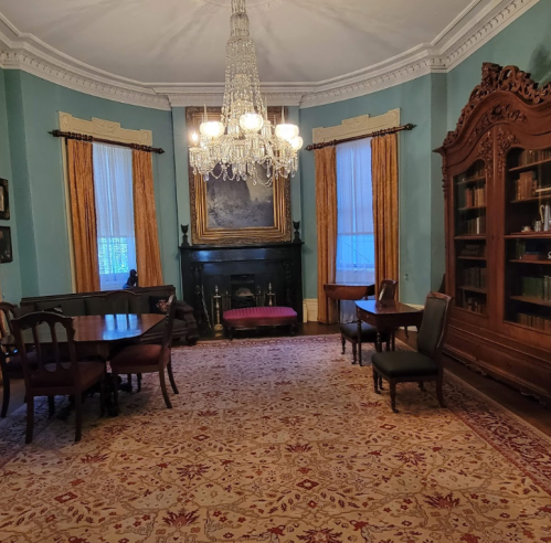 A spacious room with a chandelier, fireplace, bookshelves, and elegant furnishings, featuring blue walls and a patterned rug.