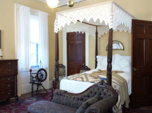 A vintage bedroom featuring a four-poster bed, ornate furniture, and a spinning wheel in a warm, inviting setting.