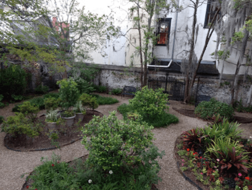 A serene garden with winding paths, lush greenery, and benches, surrounded by a historic building.