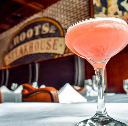 A pink cocktail in a glass, with a steakhouse sign and elegant dining setting in the background.