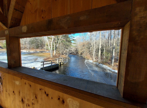 View of a serene river through a wooden structure, surrounded by trees and a hint of ice on the water.
