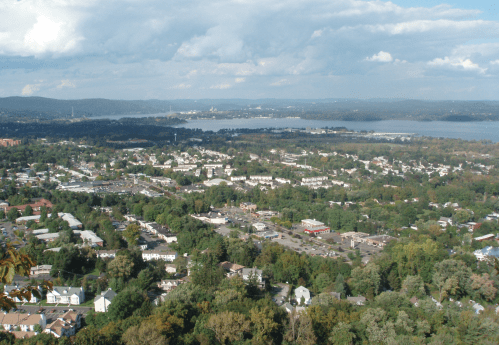 Aerial view of a town surrounded by greenery and a lake, with clouds in the sky and distant hills.
