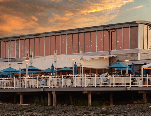 A waterfront restaurant with outdoor seating under blue umbrellas, set against a colorful sunset sky.