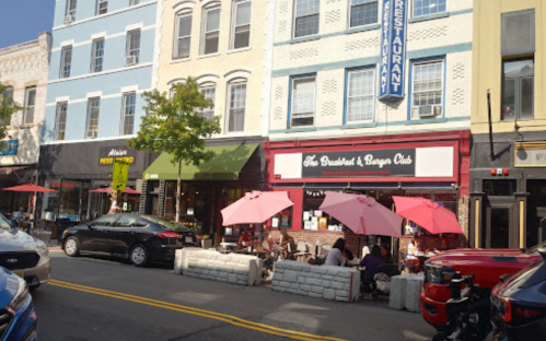 Outdoor dining scene at a restaurant with pink umbrellas, surrounded by shops and parked cars on a sunny day.
