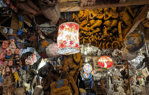 A cluttered art space filled with masks, sculptures, and colorful lanterns, featuring a mural of figures on the ceiling.