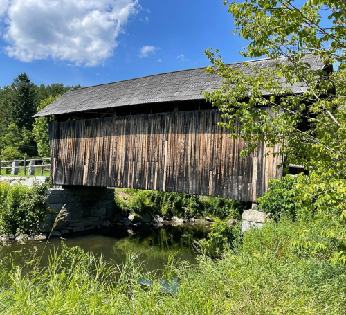 A rustic covered bridge spans a calm stream, surrounded by lush greenery and a bright blue sky.