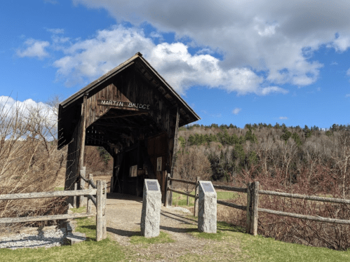 A wooden covered bridge with a sign, surrounded by trees and a blue sky with clouds. Stone markers are nearby.