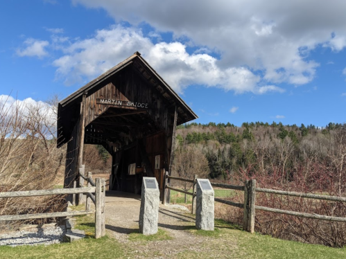 A wooden covered bridge with a sign, surrounded by trees and a blue sky with clouds. Stone markers are nearby.