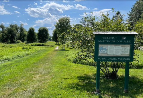 A park entrance sign welcomes visitors to Martin Bridge Park, surrounded by green grass and trees under a blue sky.