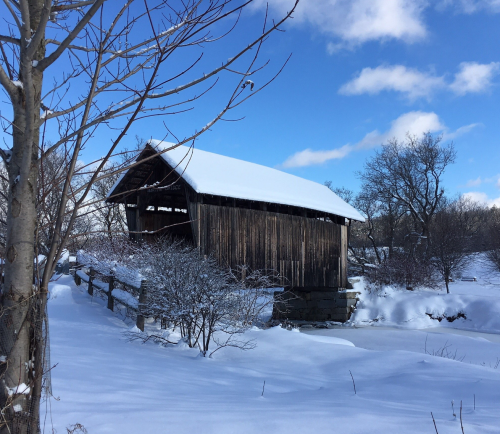 A snow-covered wooden bridge stands beside a frozen pond, surrounded by trees under a blue sky with fluffy clouds.