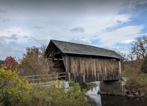 A rustic covered bridge spans a calm stream, surrounded by trees and a cloudy sky.