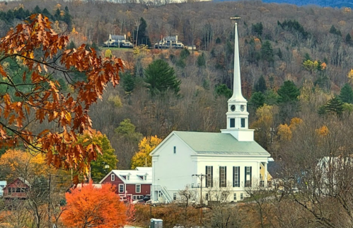 A white church with a tall steeple surrounded by autumn trees and hills in the background.
