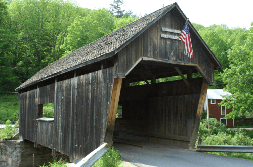 A rustic wooden covered bridge with an American flag, surrounded by lush green trees and a peaceful landscape.