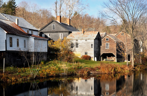 A serene scene of rustic houses by a calm pond, surrounded by autumn foliage and bare trees.