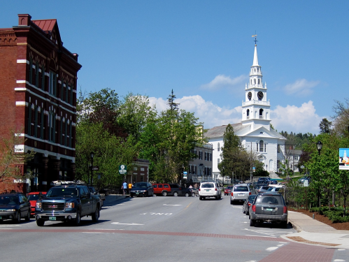 A scenic view of a small town street with a church steeple in the background and parked cars along the road.