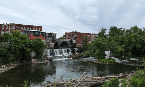 A scenic view of a river with a waterfall, surrounded by greenery and historic buildings in the background.