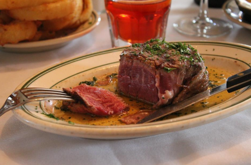 A plated steak with herbs, accompanied by a fork and knife, and a glass of drink in the background.