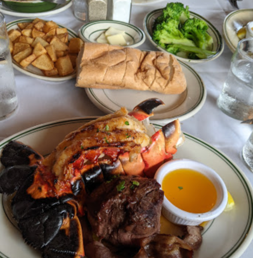 A plate featuring lobster tail, steak, butter sauce, and sides of potatoes, broccoli, and bread on a restaurant table.