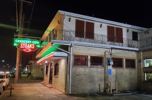 A brightly lit restaurant sign for "Crescent City Steaks" at night, showcasing a brick building with decorative railings.