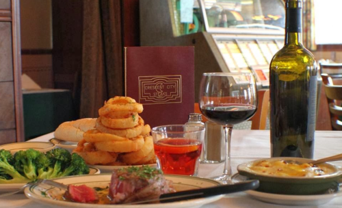 A table set with a menu, wine, a glass of red, onion rings, steak, broccoli, and a dessert in a cozy restaurant.
