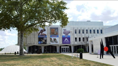 Exterior view of a museum with banners for science and history, featuring a large tree and a sculpture in the foreground.
