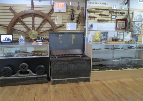 A display of historical maritime artifacts, including a ship's wheel, tools, and a chest, in a museum setting.