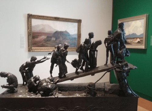 A bronze sculpture depicting a group of people crossing a bridge, set against a gallery backdrop with paintings.