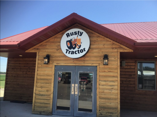 Exterior of a rustic wooden building with a red roof, featuring a sign that reads "Rusty Tractor." Clear blue sky above.