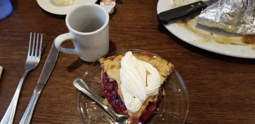 A slice of cherry pie topped with whipped cream on a glass plate, next to a coffee cup and utensils on a wooden table.