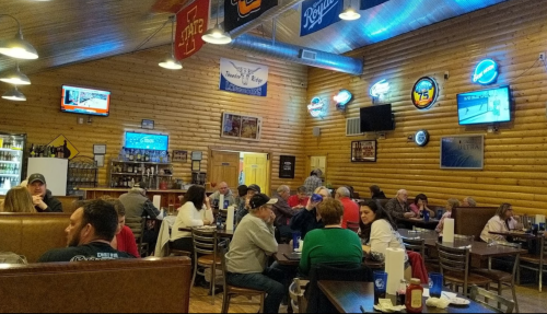 A busy restaurant interior with wooden walls, patrons dining, and various neon signs and TVs on display.