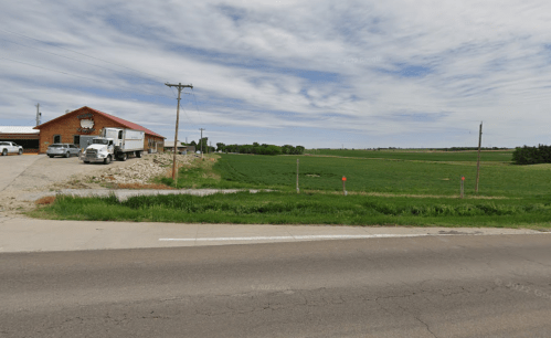 A rural landscape featuring a farm building, trucks, and expansive green fields under a cloudy sky.