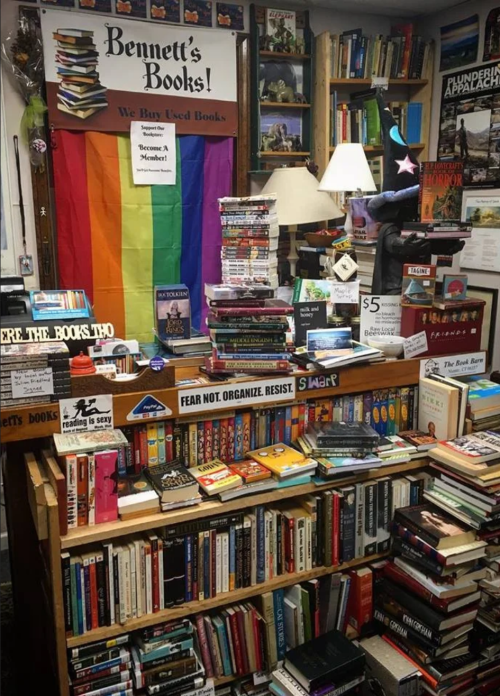 A cozy bookstore filled with stacked books, a rainbow flag, and various signs promoting reading and community engagement.