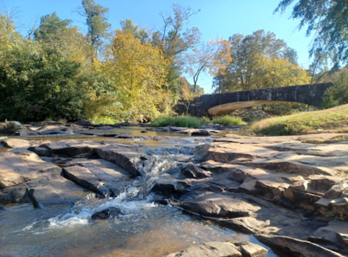 A serene landscape featuring a flowing stream, rocky shore, and a wooden bridge surrounded by autumn trees.