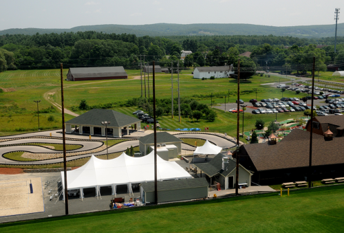 Aerial view of a go-kart track, tents, and parking lot surrounded by green fields and distant hills.