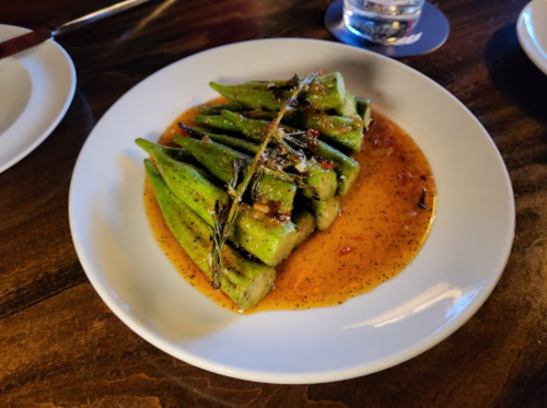 A plate of cooked okra drizzled with sauce and garnished with herbs, served on a wooden table.